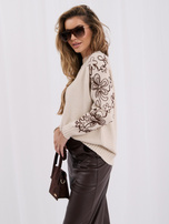 Šviesiai smėlinis moteriškas megztinis su kontrastinga siuvinėjimu.
